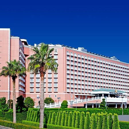 Grand Nikko Tokyo Bay Maihama Hotell Urayasu Exteriör bild The image shows a large, multi-story hotel building featuring a light pink facade. The architecture appears modern, with numerous windows arranged in a grid pattern. In the foreground, there are neatly trimmed shrubs and palm trees, adding a tropical