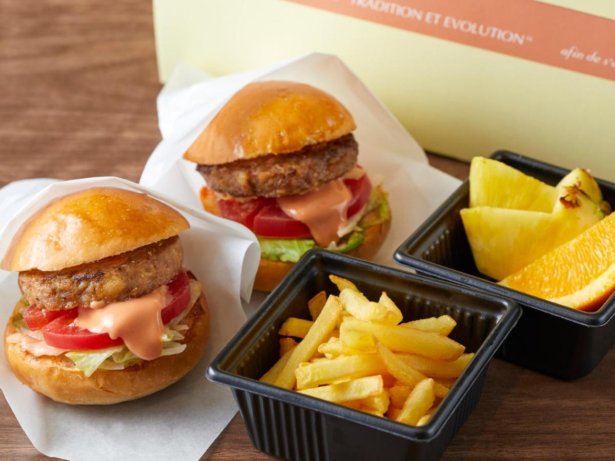 Grand Nikko Tokyo Bay Maihama Hotell Urayasu Exteriör bild The photo shows two small burgers placed on a white background, each with a golden-brown bun, a fried patty, lettuce, tomato, and a creamy sauce. Alongside the burgers, there are two containers: one holding a serving of french fries and the other con