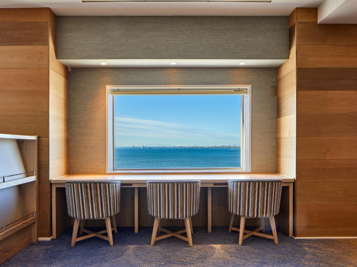 Grand Nikko Tokyo Bay Maihama Hotell Urayasu Exteriör bild The photo shows a modern workspace with three striped chairs positioned at a long, white desk. The desk faces a large window with a view of a tranquil body of water and a distant skyline. The walls around the workspace are made of wood paneling, crea