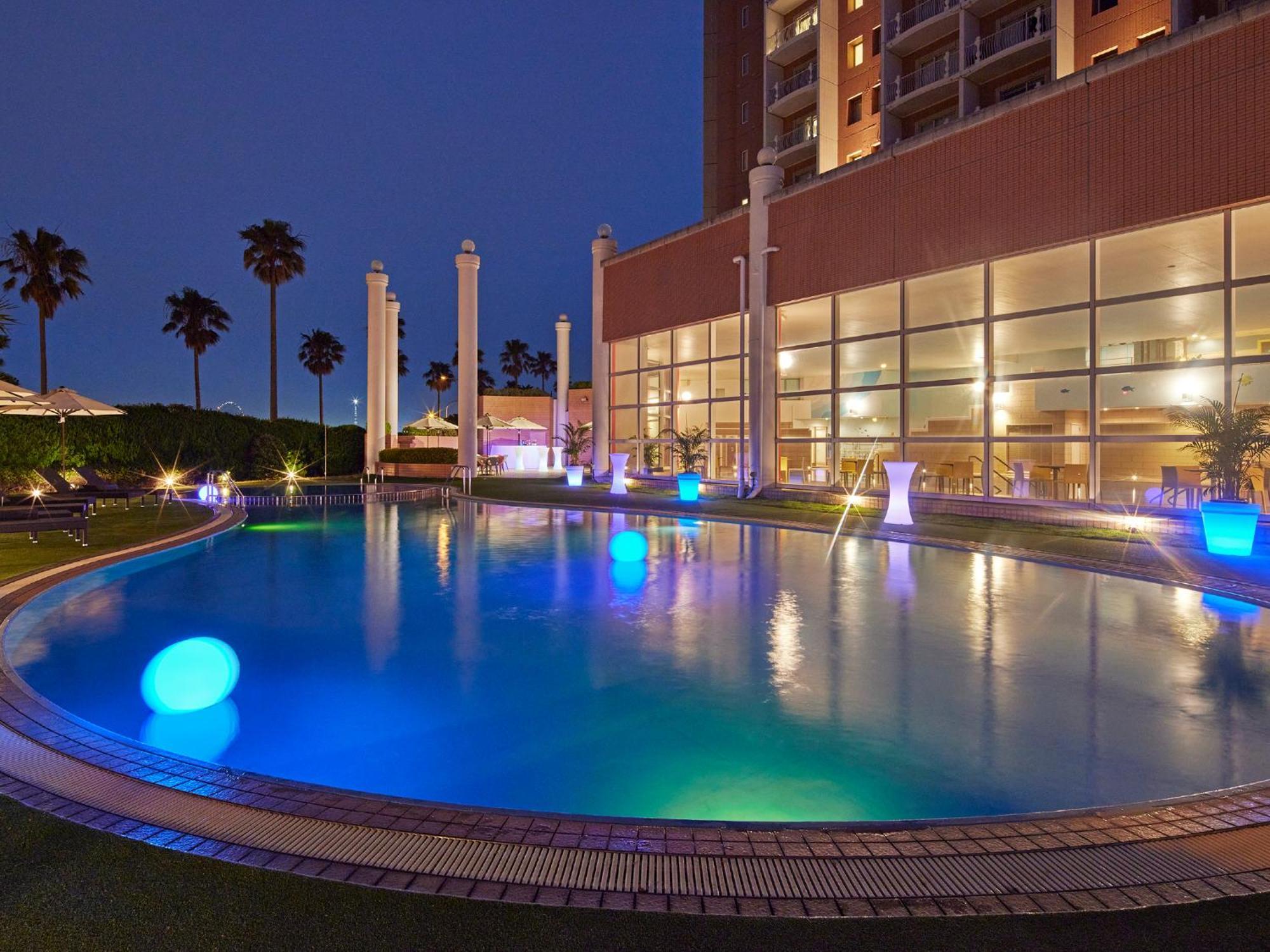Grand Nikko Tokyo Bay Maihama Hotell Urayasu Exteriör bild The photo shows a well-lit swimming pool at night, surrounded by a lush green area. The pool features glowing blue lights that create a serene and inviting atmosphere. Palm trees can be seen in the background, adding a tropical feel to the scene. Add