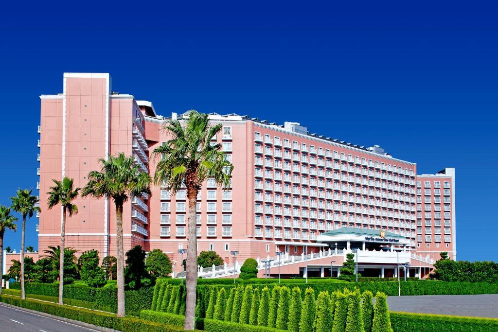 Grand Nikko Tokyo Bay Maihama Hotell Urayasu Exteriör bild The image shows a large, multi-story hotel building featuring a light pink facade. The architecture appears modern, with numerous windows arranged in a grid pattern. In the foreground, there are neatly trimmed shrubs and palm trees, adding a tropical
