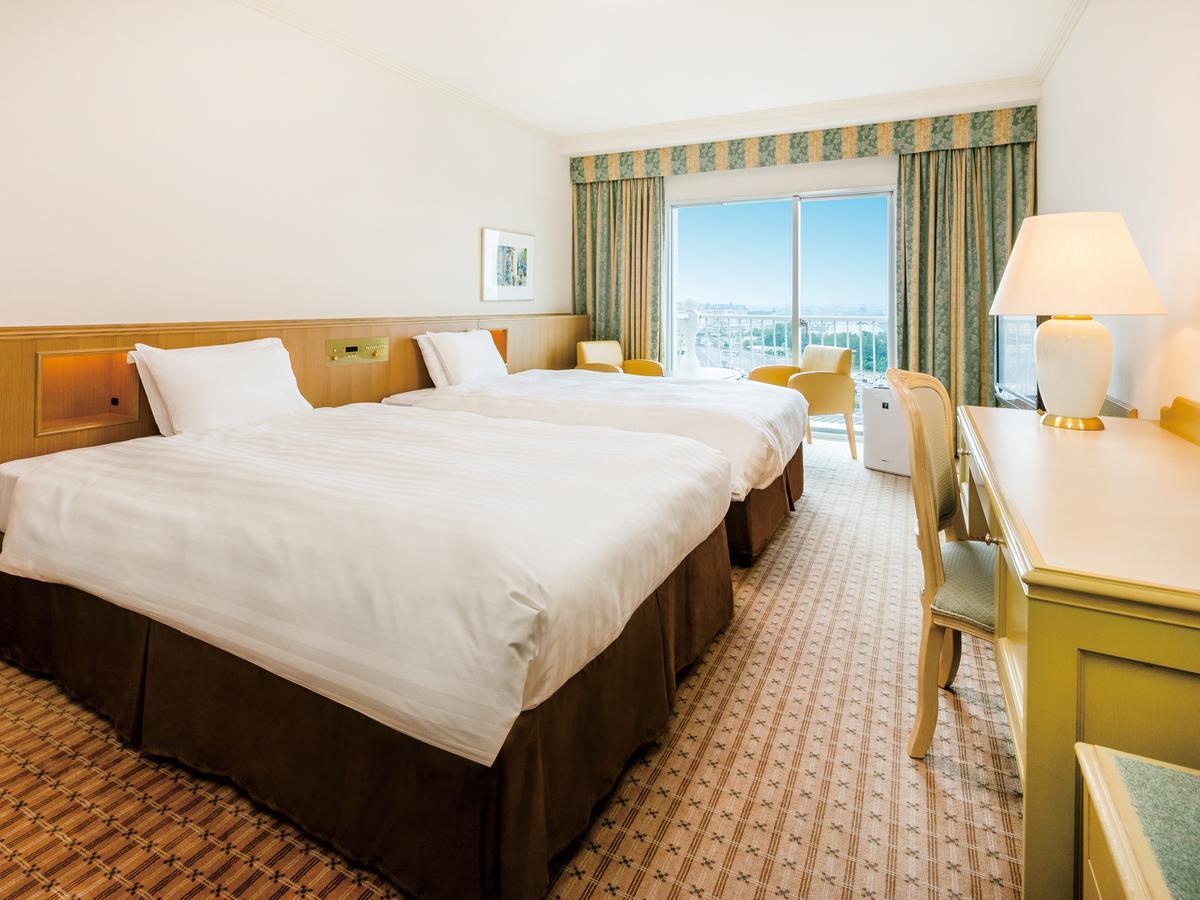 Grand Nikko Tokyo Bay Maihama Hotell Urayasu Exteriör bild The photo shows a hotel room featuring two twin beds with white linens and dark brown bed skirts. The room has a light, neutral-colored wall and carpet. There is a window with curtains that let in natural light, offering a view outside. A small desk 